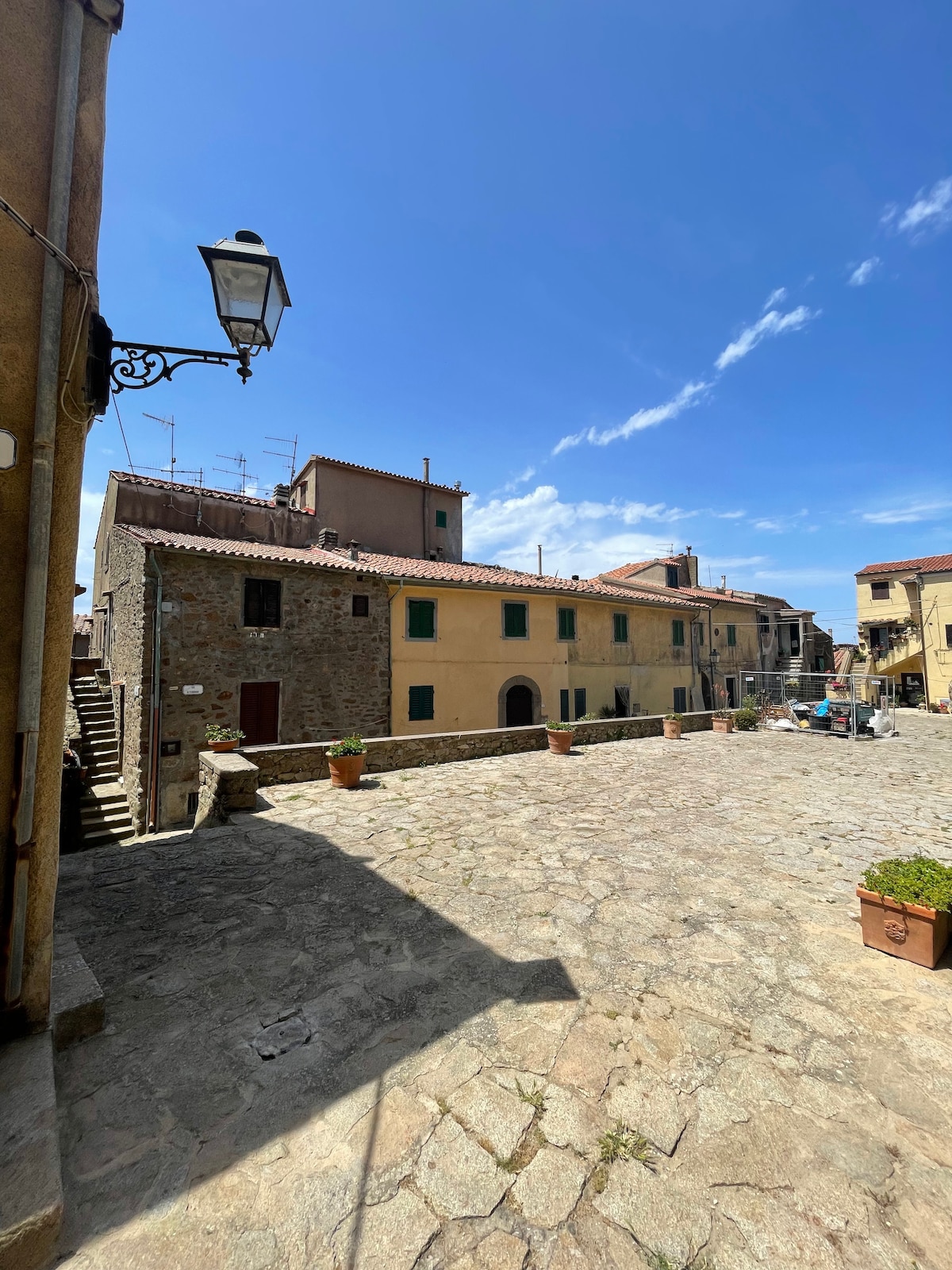 A cobblestone square is shown, bordered by rustic buildings featuring a mix of stone and yellow facades. Potted plants add greenery to the scene, under a clear blue sky. A few chairs and tables are visible, suggesting an inviting space for relaxation.