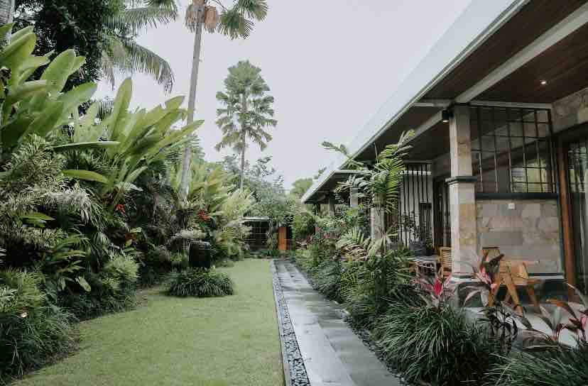 A lush garden is set alongside a well-maintained walkway, surrounded by vibrant greenery and tropical plants. The outdoor area features stone pathways leading to a welcoming entrance, with wooden chairs positioned to offer a serene view of the natural surroundings.