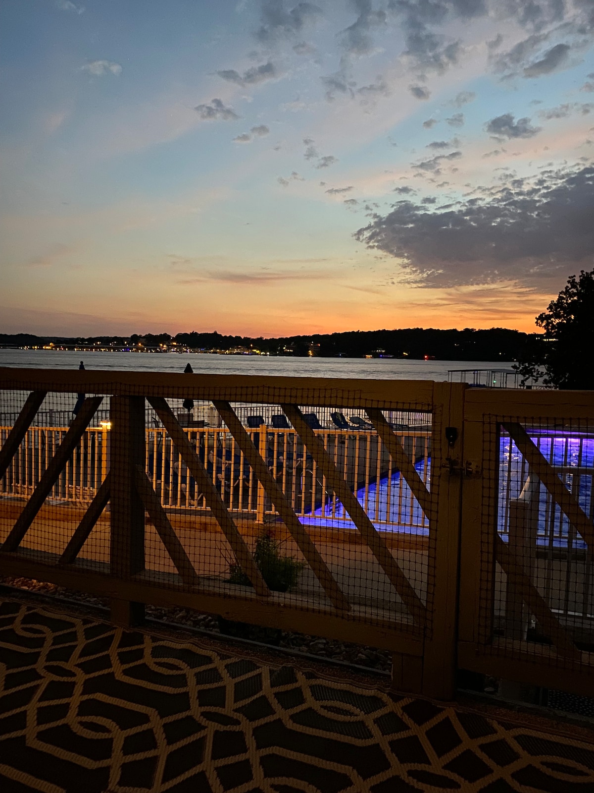 A sunset view is captured from the patio, showcasing a lake surrounded by hills. Soft clouds are visible in the evening sky, while the pool area is illuminated below, reflecting subtle hues of blue. A railing provides a barrier while the patio flooring features a patterned design.