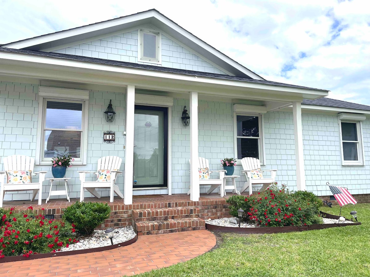 A charming exterior view of a cottage features a welcoming porch with two white chairs adorned with colorful cushions. Potted flowers are placed near the entrance, with neatly arranged bushes and a small American flag enhancing the tidy landscape.
