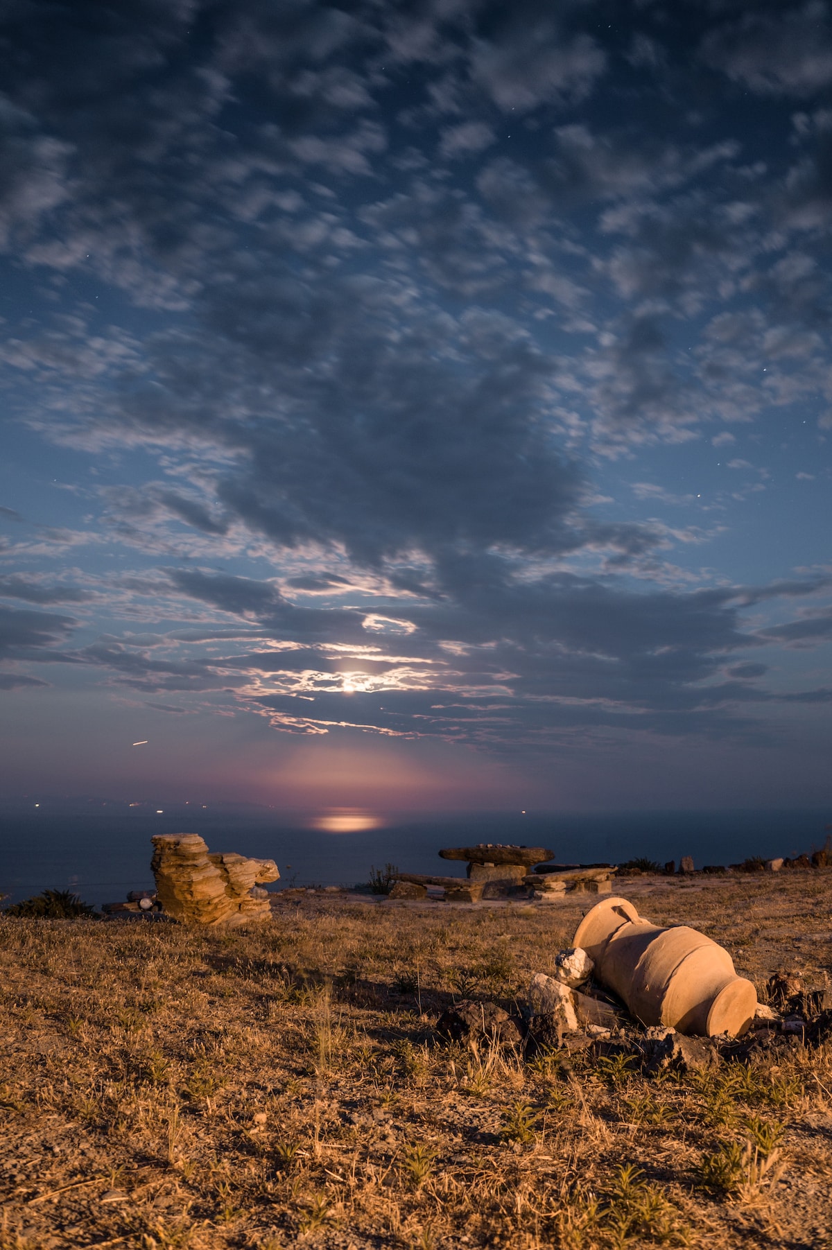 A serene landscape is observed under a night sky with scattered clouds. The moonlight reflects on the sea, illuminating the horizon. In the foreground, rocky formations are positioned beside patches of grass, creating a peaceful atmosphere that invites reflection and quiet appreciation of nature.