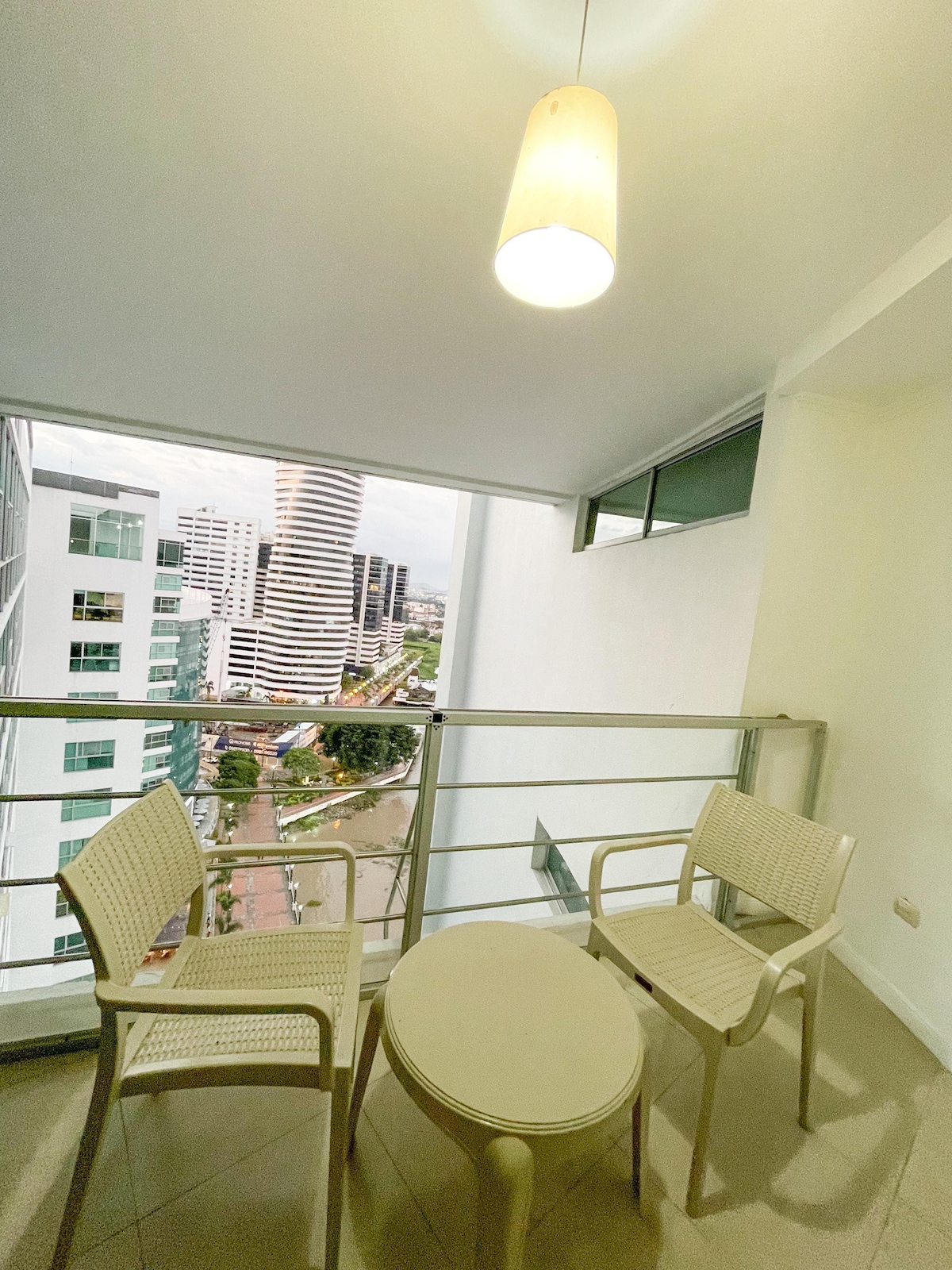 A small balcony area is presented, featuring two light-colored chairs and a round table. A large window provides a view of the surrounding cityscape. A pendant light hangs above, adding a touch of illumination to the space.