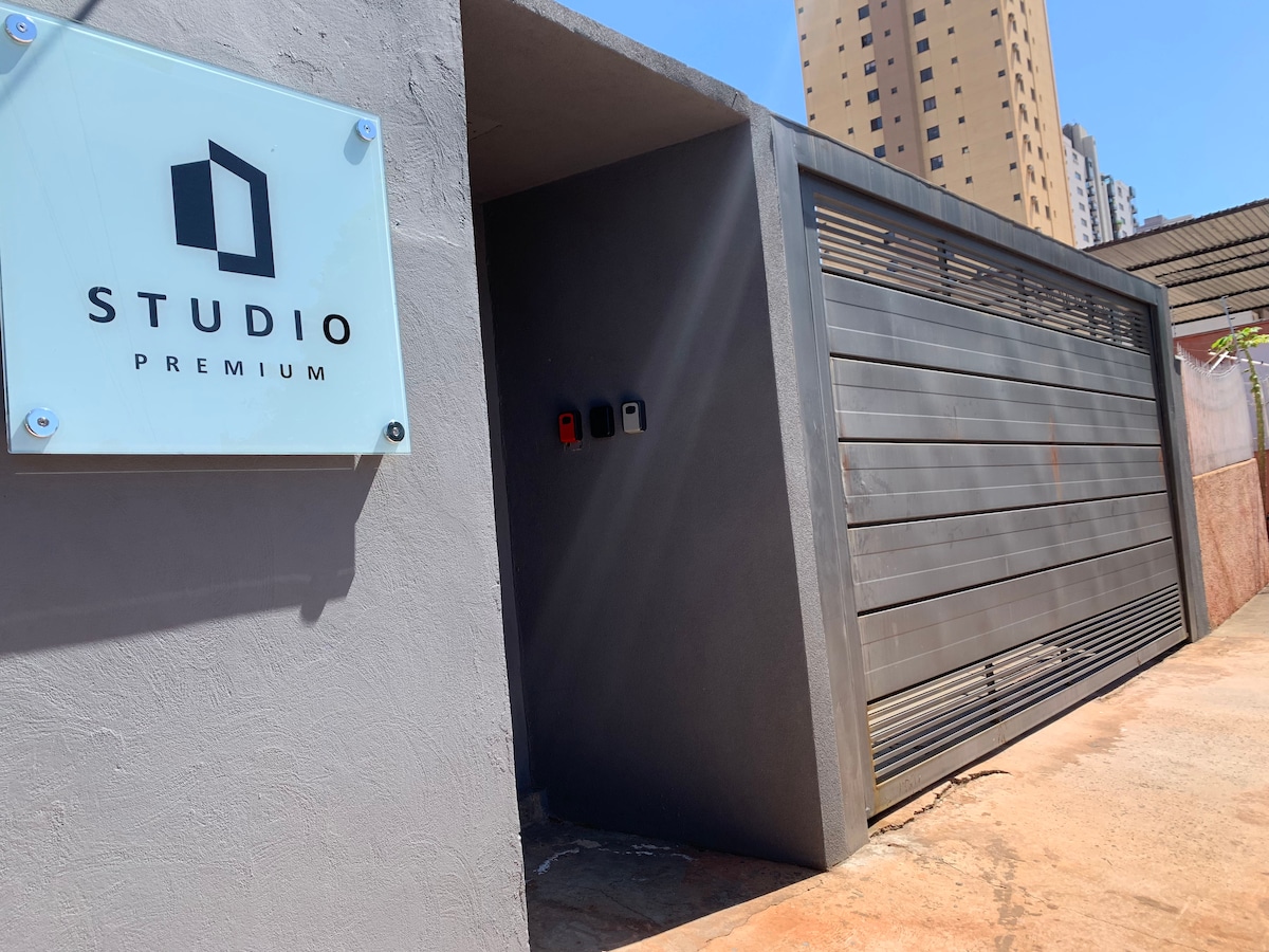 The exterior entrance of STUDIO PREMIUM 2 features a modern sign, positioned alongside a sleek, closed garage door. Sunlight reflects off the surface, creating a sense of brightness. Surrounding buildings are partially visible in the background, indicating an urban setting.