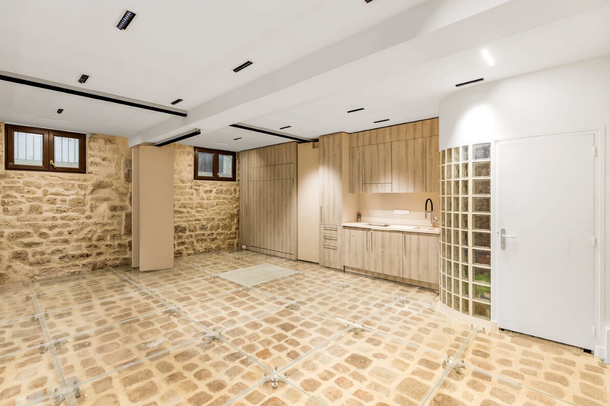 A spacious interior showcases exposed stone walls and a distinctive glass floor revealing 16th-century cobblestones beneath. An equipped kitchen is visible, with modern cabinetry and a sleek countertop. Natural light enters through small windows, enhancing the open layout suitable for various activities.