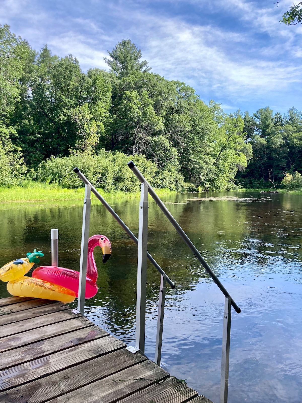 A wooden dock extends into a calm river, featuring a colorful inflatable flamingo and pineapple floating nearby. Lush greenery lines the riverbank, reflecting softly on the water's surface under a bright blue sky, creating a serene outdoor environment.
