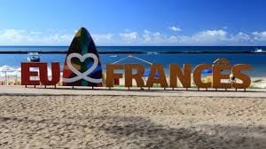A large colorful sign reading 'EU ♥ FRANcÊS' is positioned on a sandy beach, facing a calm blue ocean. The clear sky above showcases a few white clouds, adding to the serene beach atmosphere.