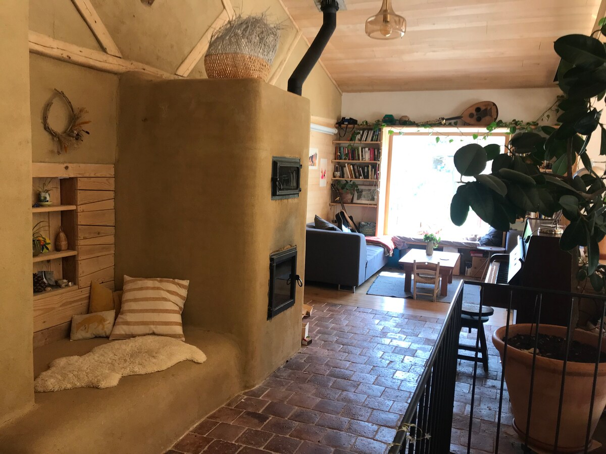 The interior features a warm, earthy wall texture with a cozy seating nook beside a wood stove. A large window allows natural light to fill the space, which includes a inviting living area with bookshelves, a sofa, and a piano visible in the background.