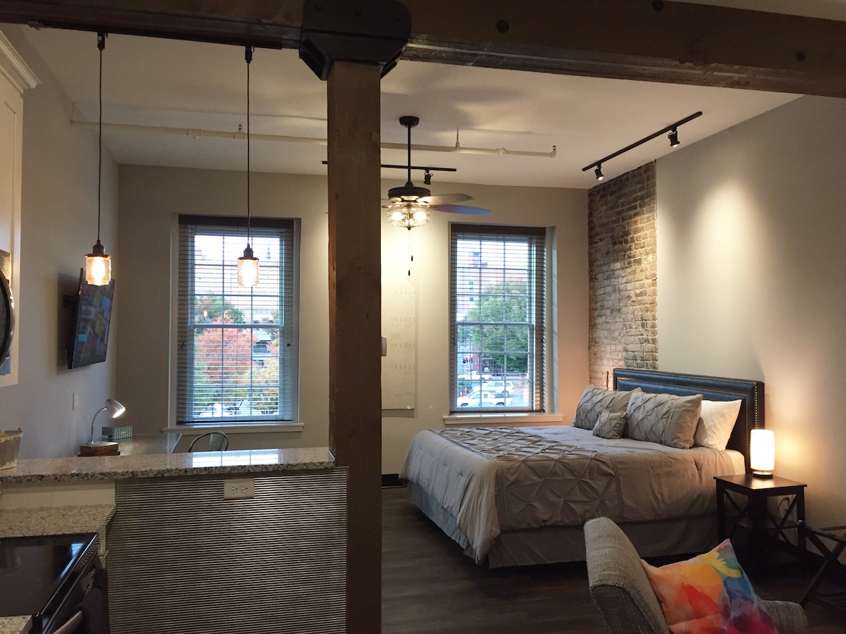 A modern bedroom features a spacious bed with soft linens, positioned near large windows that provide natural light. A contrasting brick wall adds texture to the space, and light fixtures create a cozy ambiance. A kitchen area is visible in the background.