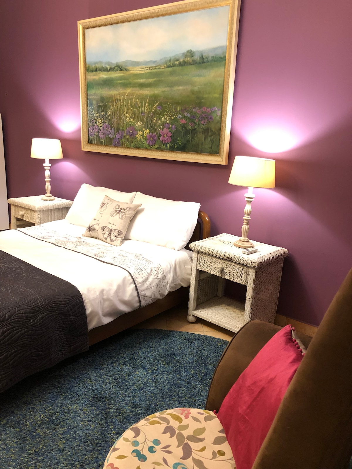 A sleeping area features a spacious bed with a decorative throw and a floral cushion. Two bedside tables with lamps are placed on either side, casting a soft glow. A colorful rug underfoot complements the warm purple walls adorned with a landscape painting.