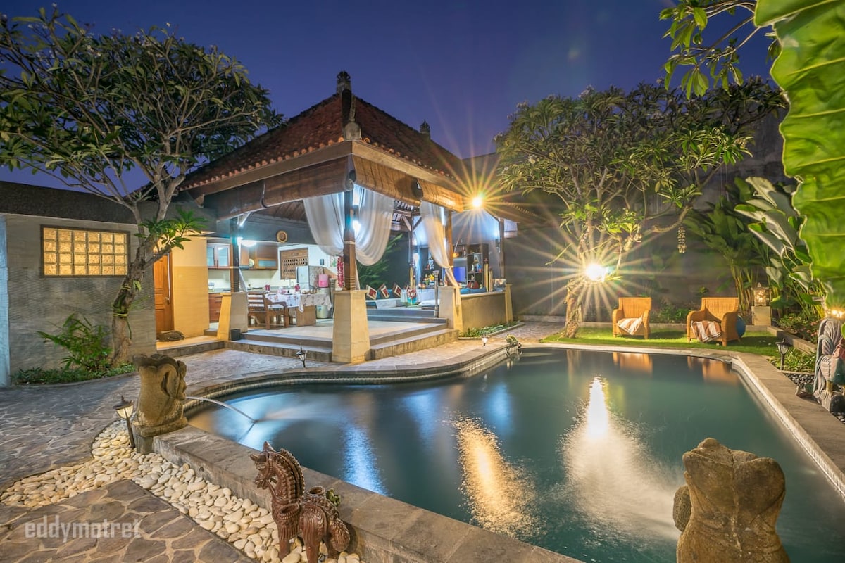 A serene villa is highlighted by a tranquil swimming pool reflecting surrounding greenery and soft lighting. The open pavilion features comfortable seating and dining areas, inviting relaxation. Lush trees and subtle sculptures enhance the outdoor space, creating a harmonious blend of nature and design.