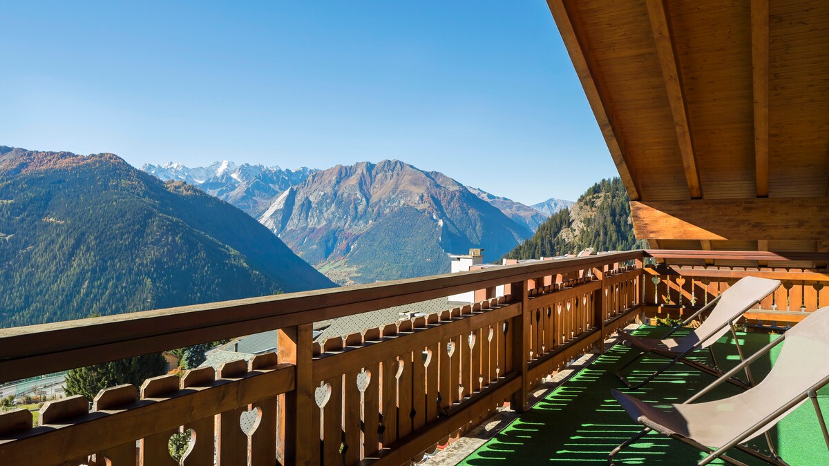 A spacious balcony is shown, featuring wooden railings with decorative cut-outs and two reclining lounge chairs positioned for relaxation. The stunning mountain landscape stretches before the viewer, with blue skies and distant peaks visible.