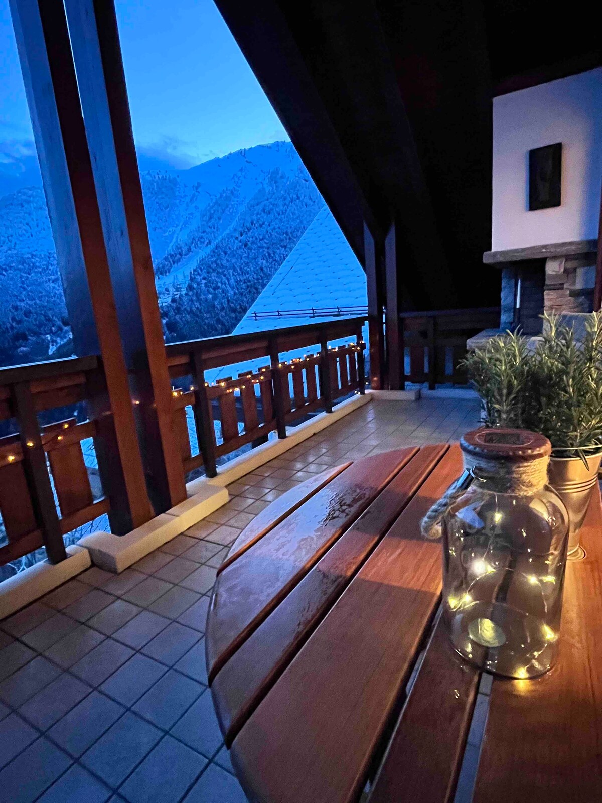 A spacious balcony is shown with wooden railings and a tiled floor. A round wooden table is present, accompanied by a glass bottle filled with fairy lights. Scenic mountain views can be seen in the background, with a soft dusk light illuminating the space.