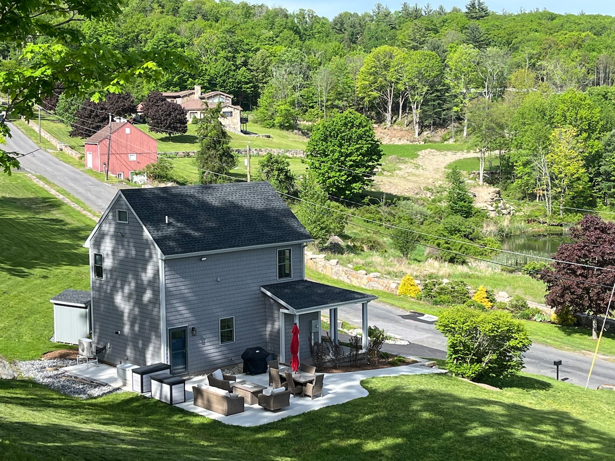 The exterior of the tiny home is showcased, set against a backdrop of lush greenery. A spacious patio area features a fire pit and seating arrangement, complemented by a red umbrella providing shade. The surrounding landscape includes trees and a glimpse of a nearby pond.