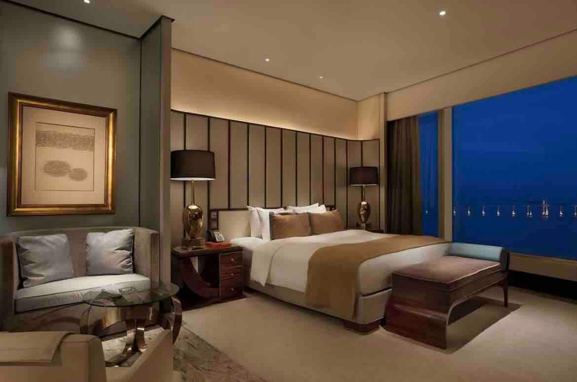 A spacious bedroom features a large bed covered with soft linens and accented by warm, neutral tones. A comfortable seating area is present, consisting of a chair and small table. Soft lighting complements the modern decor, with large windows offering views of the nighttime skyline.