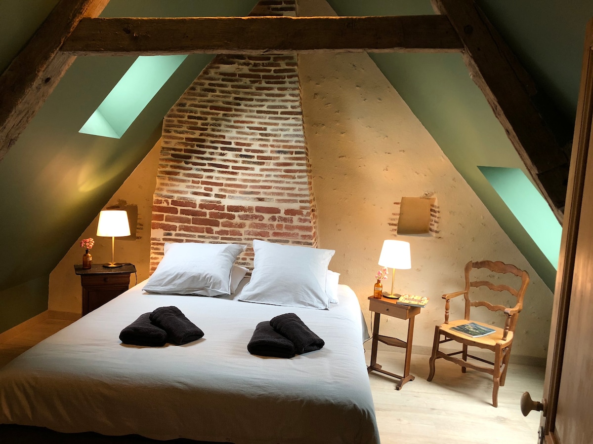 A cozy bedroom with sloped ceilings features a spacious bed adorned with soft linens. Two bedside tables with lamps provide gentle illumination. A wooden chair is positioned nearby, creating a warm and inviting retreat. Exposed brick and wooden beams highlight the rustic charm.