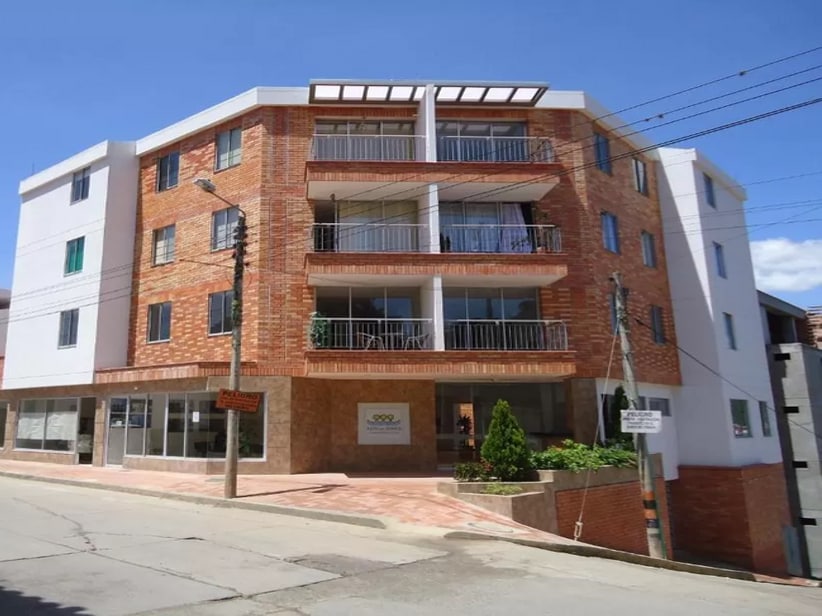 An exterior view of a modern four-story apartment building is presented. The facade features a combination of brick and white finishes, with multiple balconies offering outdoor space. Ground-level greenery and signage are visible, providing a welcoming atmosphere for residents and visitors.