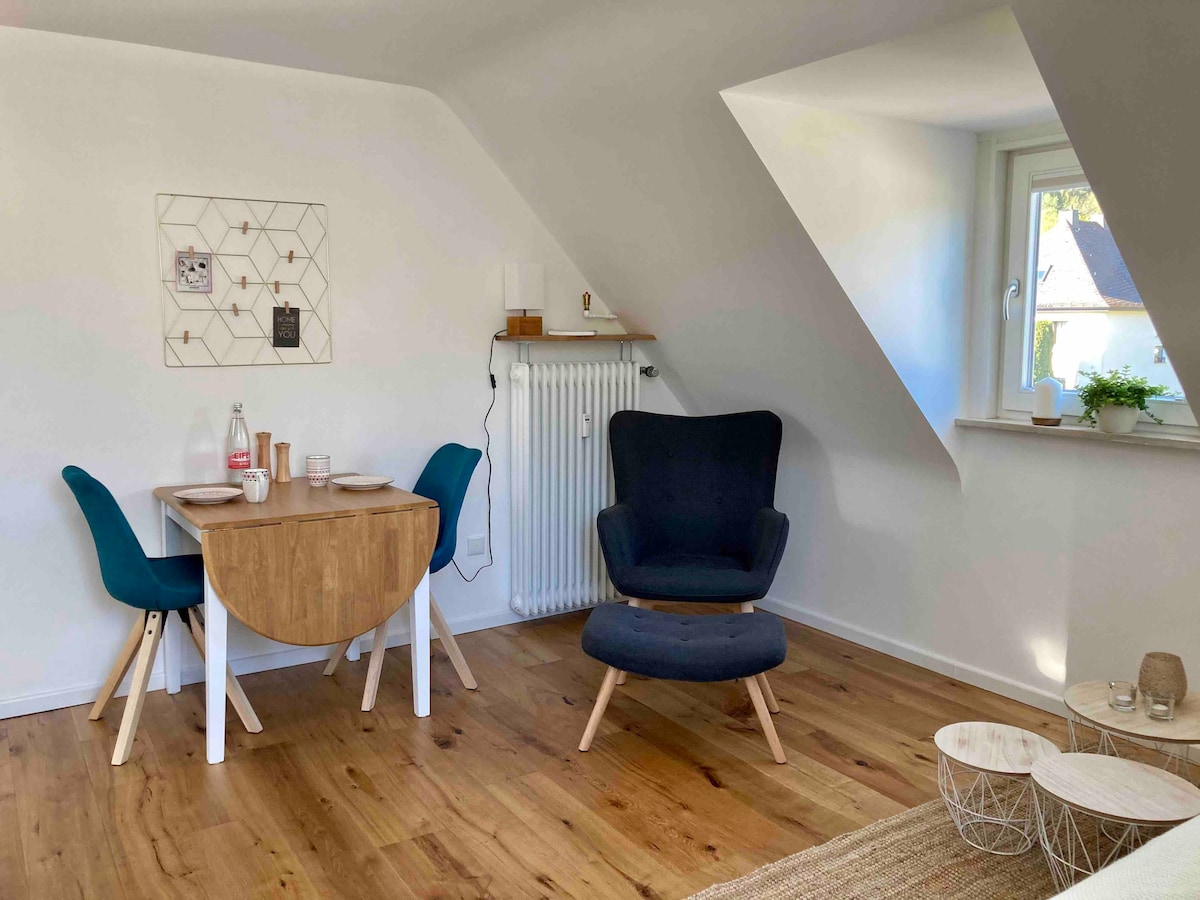 A cozy corner of the apartment features a round, wooden dining table with two blue chairs set for a meal. A comfortable, dark chair is positioned nearby. Soft natural light enters through a small window, highlighting the wooden floor and bright, uncluttered walls.