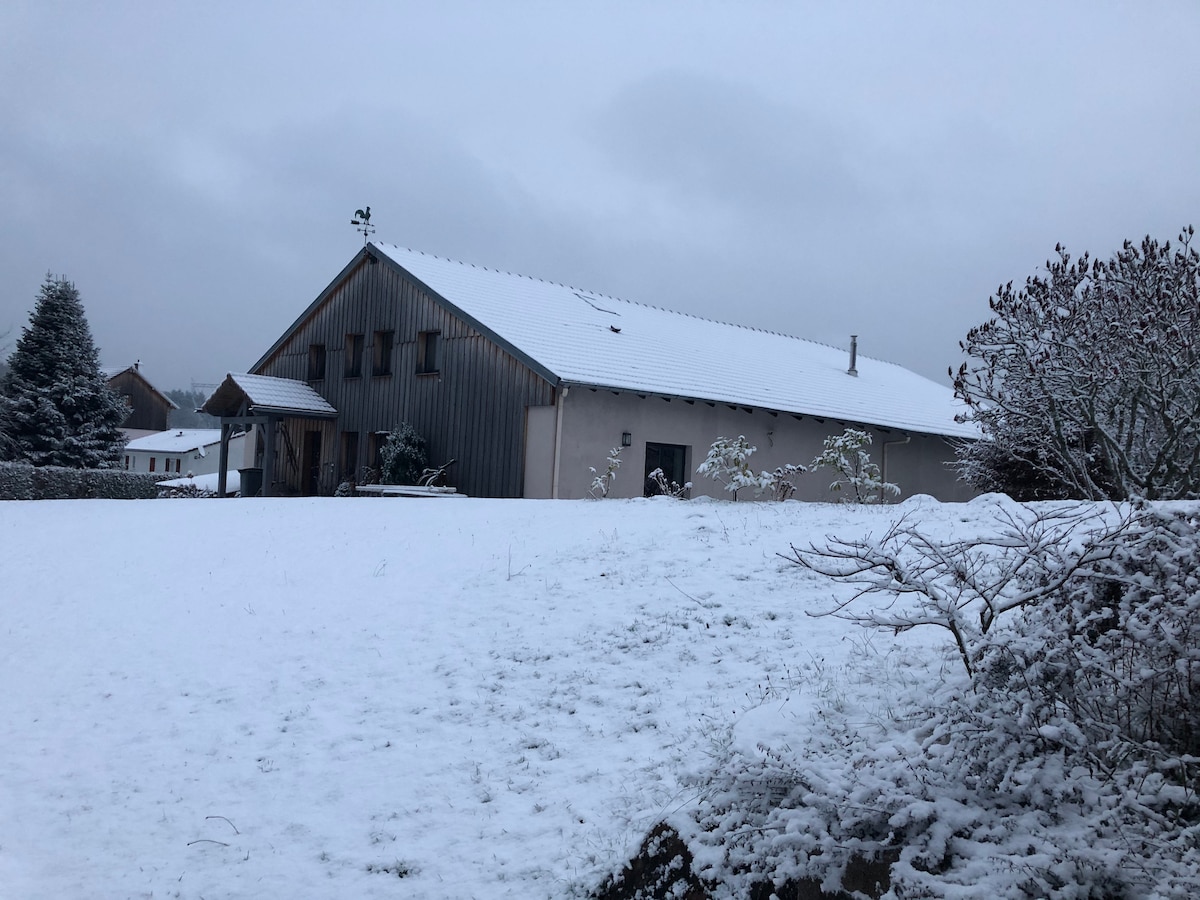 A spacious farmhouse is nestled in a snowy landscape, with a sloped roof and a mix of wooden and metal siding. Fresh snow blankets the surrounding ground and trees, creating a serene winter scene under a cloudy sky.