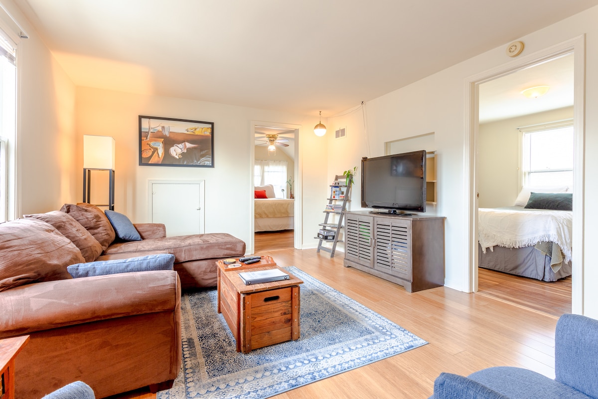 A cozy living area features a brown sectional sofa accompanied by a small wooden coffee table. A flat-screen TV is positioned on a side cabinet. Light-colored walls and hardwood flooring enhance the bright ambience, while two doorways lead to separate bedrooms.