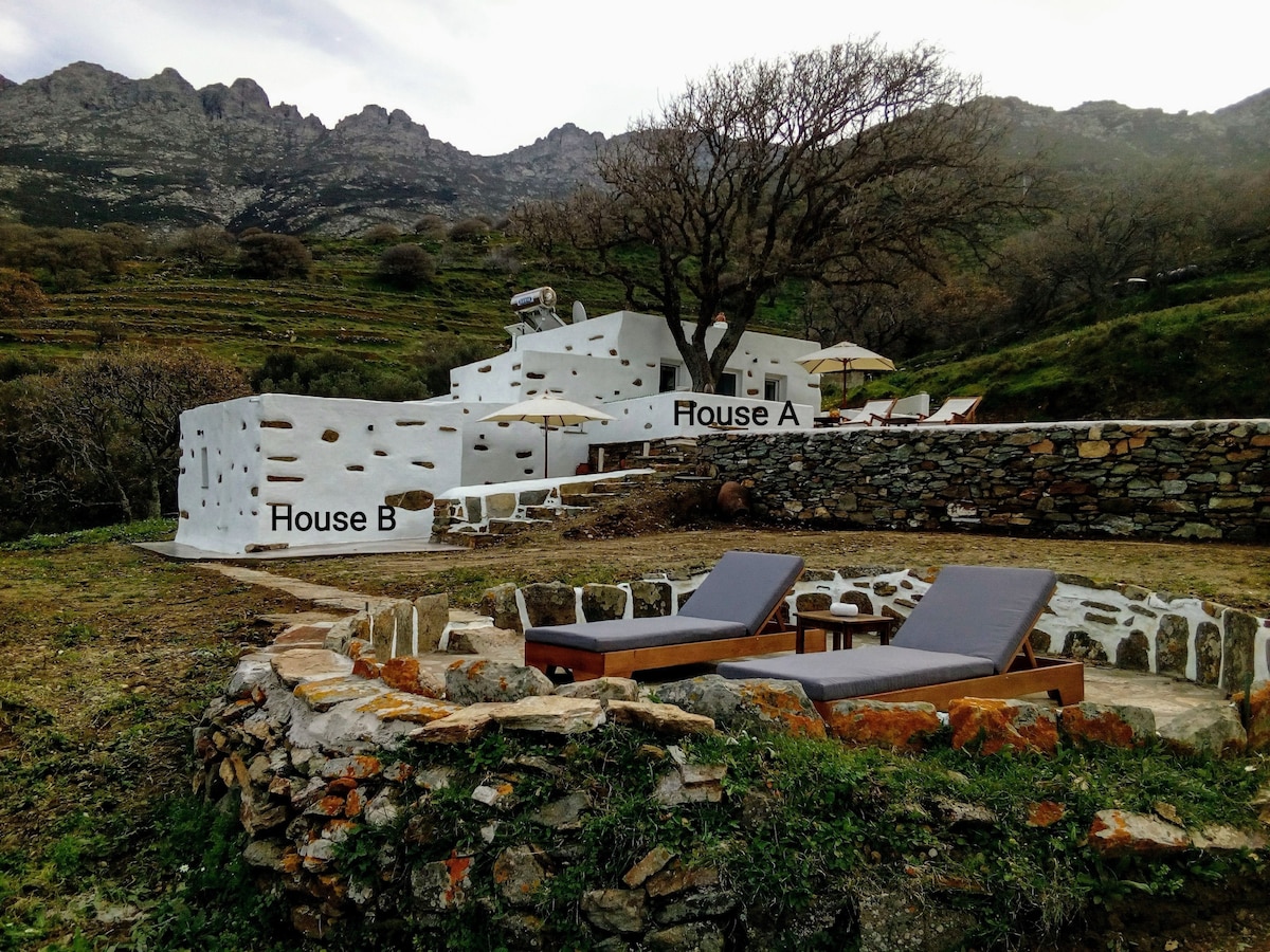 The image displays two stone-accented white houses, labeled House A and House B, nestled in a scenic, grassy area beneath mountainous terrain. Simple lounge chairs are positioned near a circular stone seating area, providing a welcoming outdoor space. Minimalist design elements are featured throughout the surroundings.