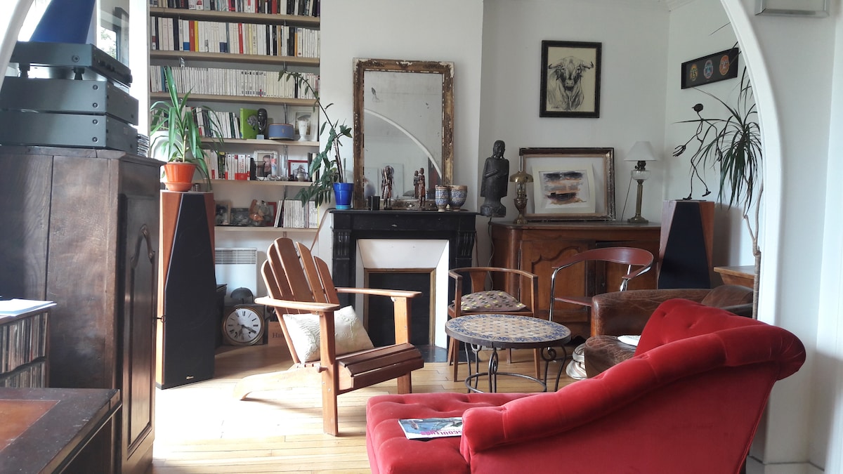 A living area is presented with a mix of seating options, including an upholstered red chair and a wooden lounge chair. Bookshelves filled with various items line the walls, while a decorative mirror and artwork add character. Natural light fills the space, highlighting wooden flooring.