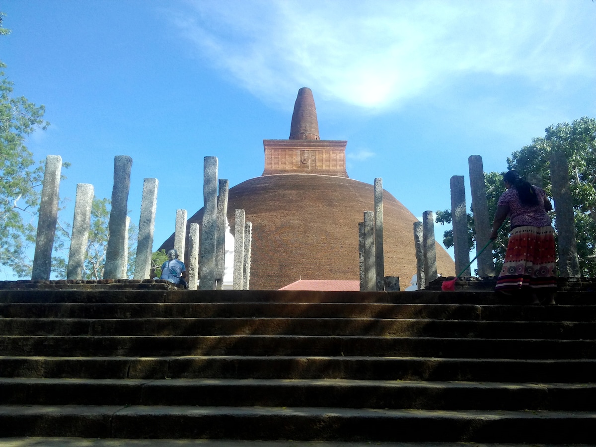 A large brick stupa rises prominently against a clear blue sky, surrounded by weathered stone pillars. Steps lead up to the structure, where visitors can be seen ascending. Lush greenery frames the scene, adding natural elements to the historical backdrop.