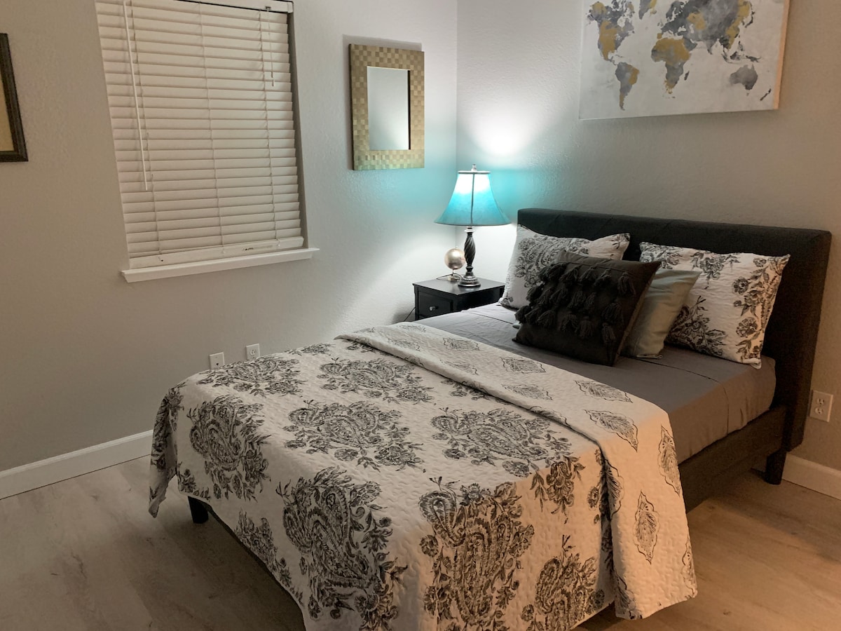 A spacious bedroom is furnished with a full-size bed covered in a decorative quilt. A bedside table with a lamp is positioned next to the bed. A world map hangs on the wall, and a window with blinds allows for natural light.