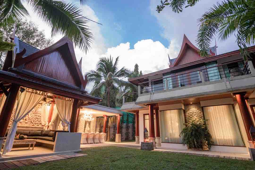 A modern villa designed in traditional Thai architecture is surrounded by lush greenery. The structure features a spacious outdoor lounge area with comfortable seating under draped fabric. The garden includes well-maintained grass and tropical plants, creating a serene and inviting atmosphere.