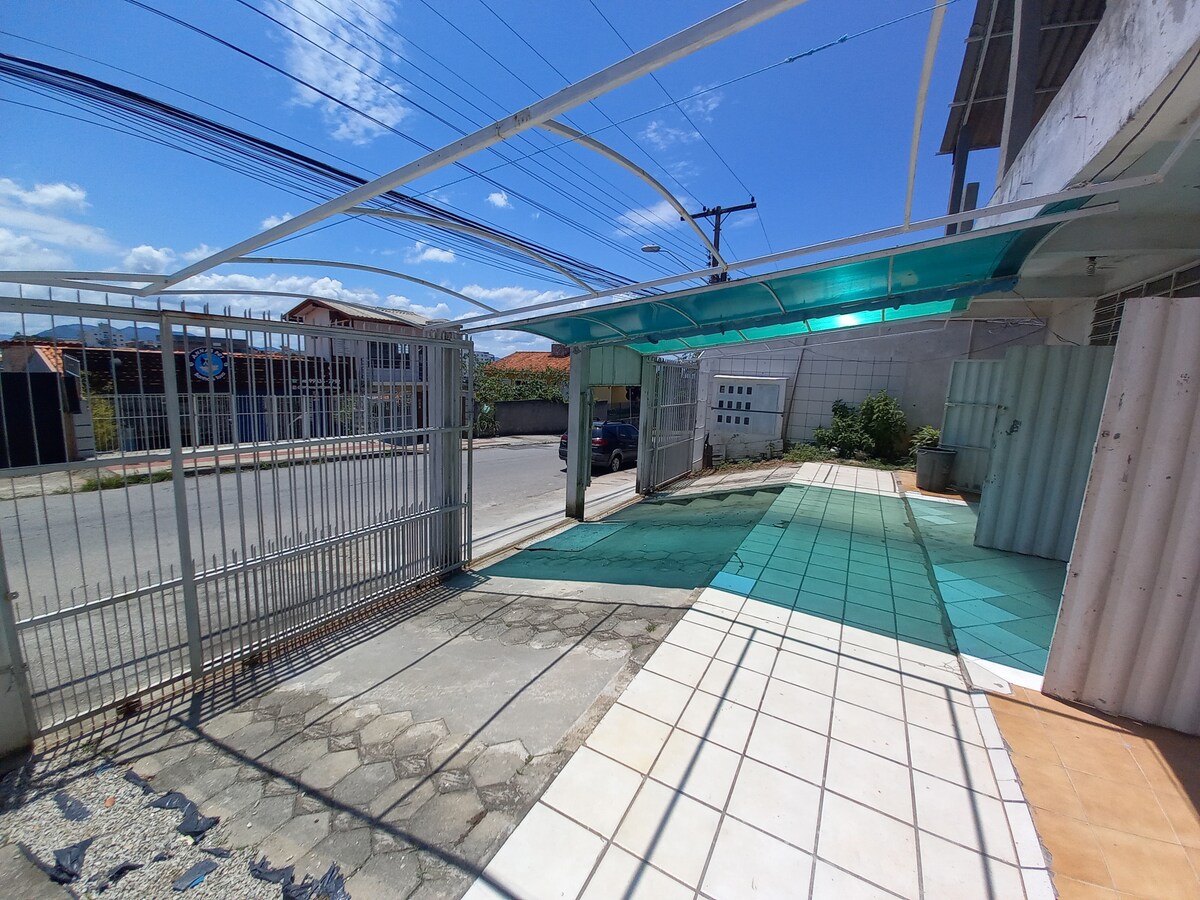 An inviting entrance showcases a spacious area with tiled flooring, bordered by low walls. A gate provides access to the street, with sunlight highlighting the blue-green canopy overhead. The surrounding area features hints of greenery and a clear blue sky.