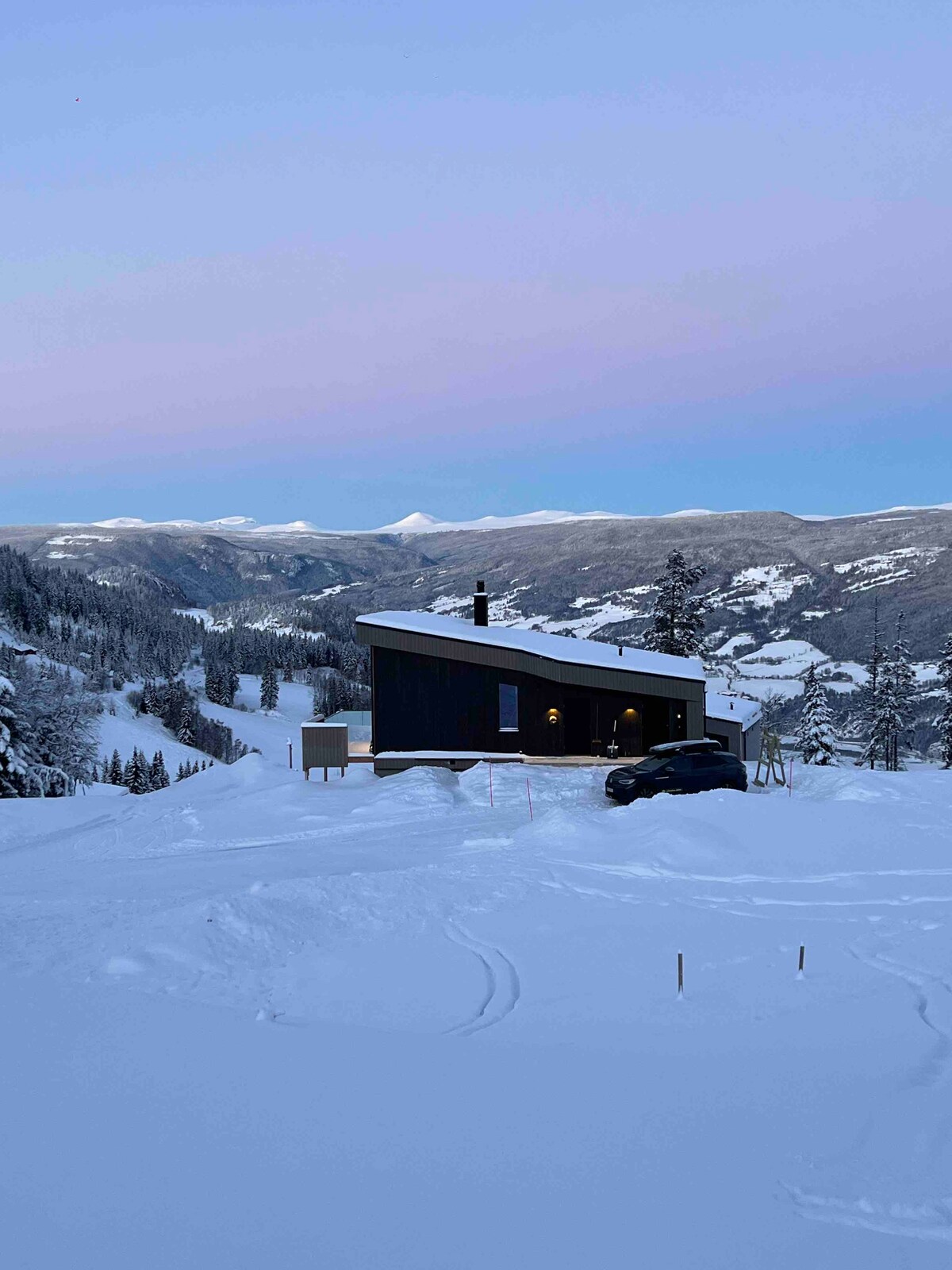A modern cabin sits nestled in a snowy landscape, surrounded by mountains. The structure features a sleek design with large windows. A vehicle is parked nearby, and the tranquil scene is enhanced by the gentle hues of the twilight sky.