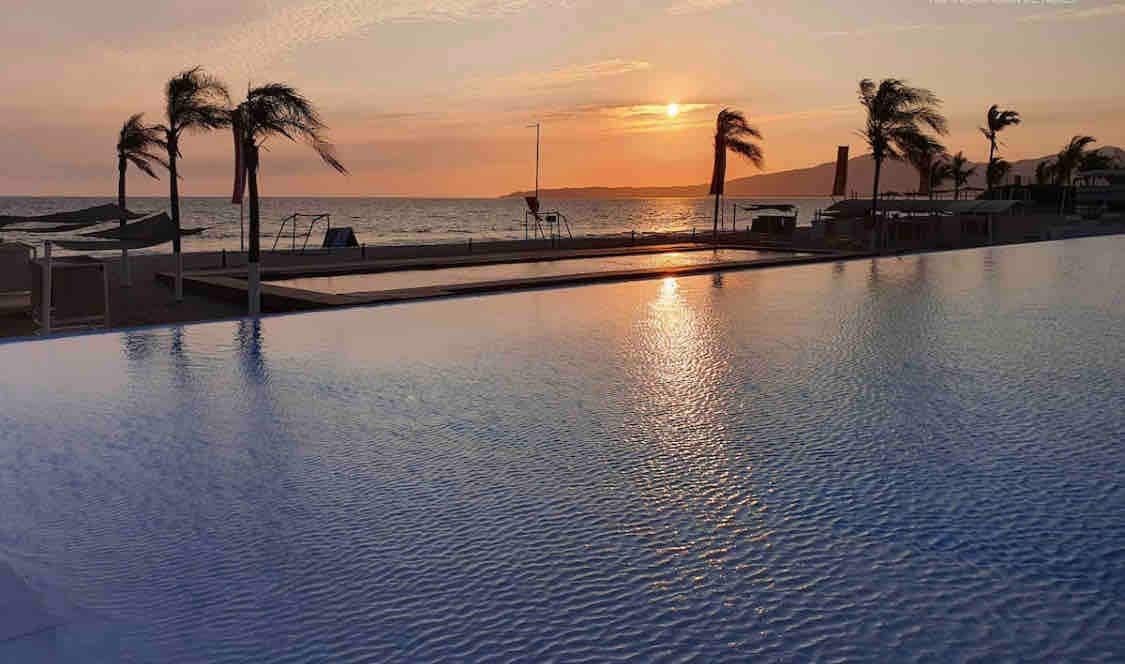 A tranquil infinity pool reflects the colors of the sunset over the ocean. Ripples extend across the water's surface, glimmering as the sun descends on the horizon. Palm trees gently sway in the evening breeze, enhancing the serene coastal ambiance.