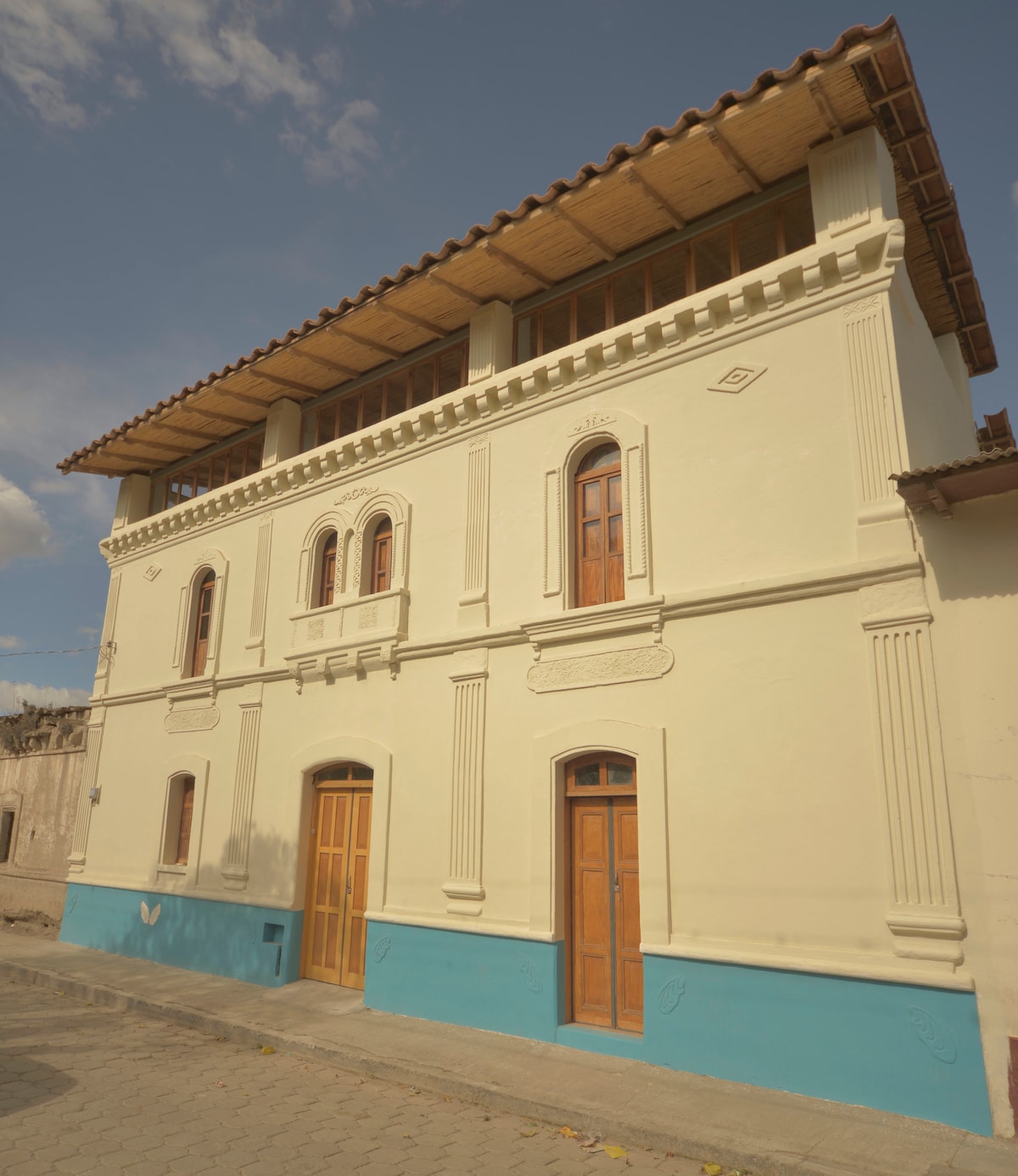 A three-story building features a light-colored façade with decorative elements and wooden doors. The top floor has a covered balcony with wooden beams, while the lower sections display contrasting blue accents. Natural light illuminates the exterior, highlighting the building’s architectural details.