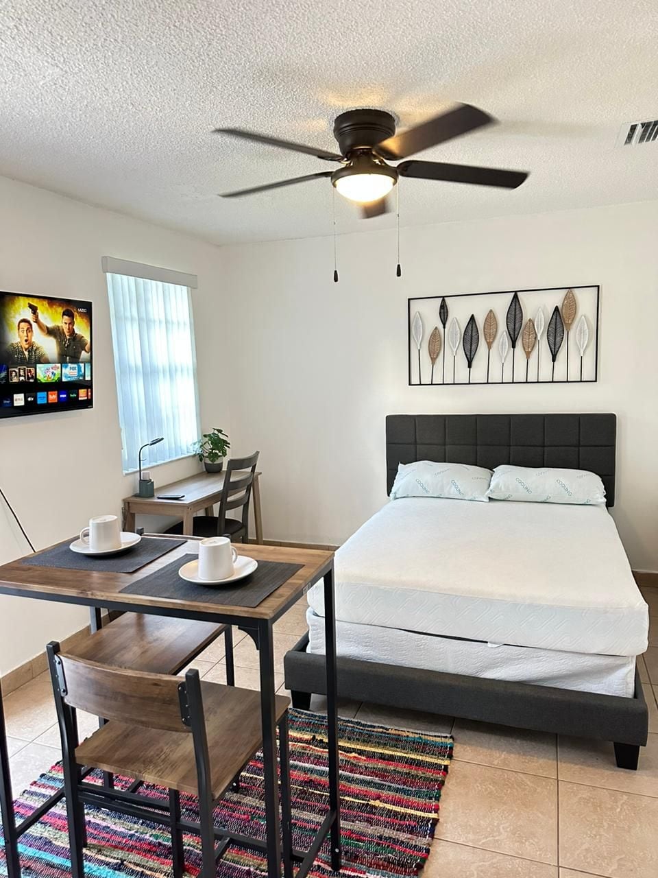 A cozy studio features a spacious bed with light bedding, complemented by a ceiling fan above. A small dining area holds two chairs and a table, while a television is mounted on the wall. Natural light filters through sheer curtains, creating a bright atmosphere.