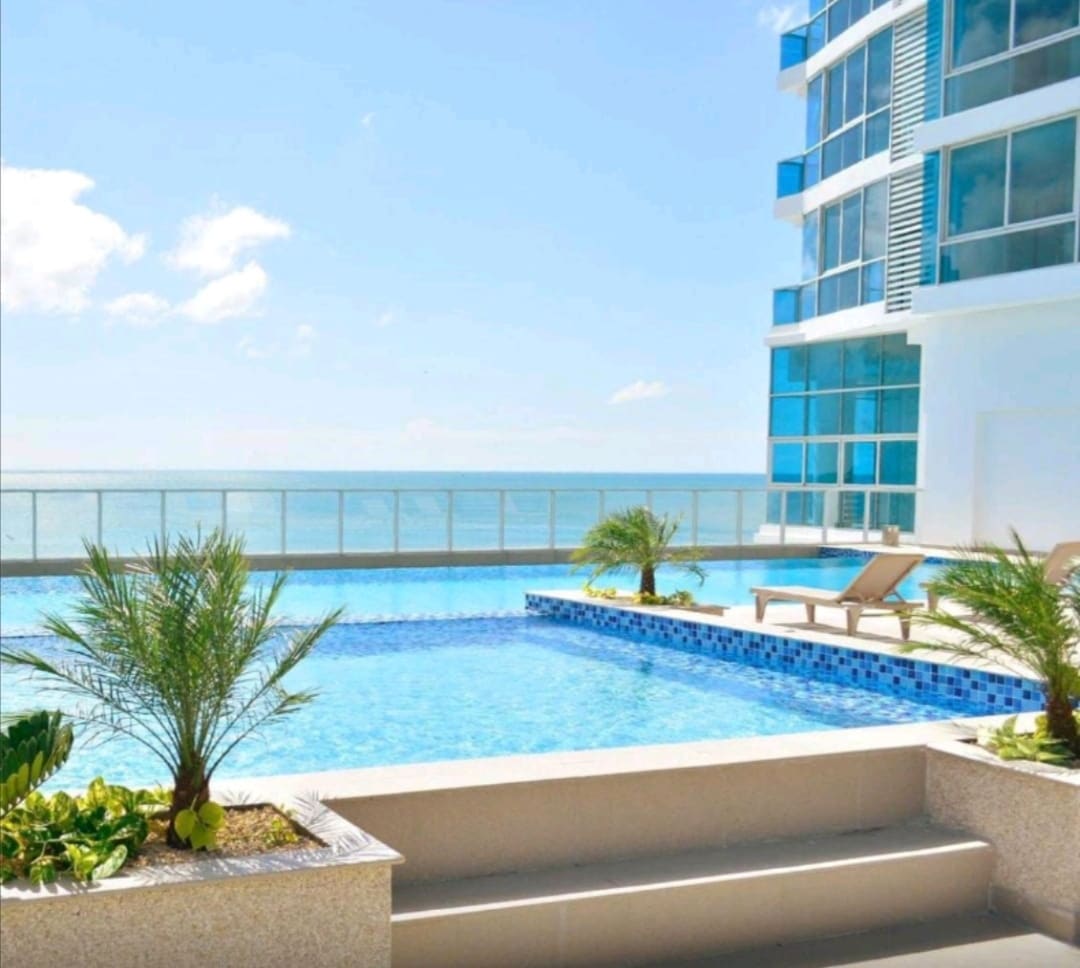 An outdoor pool is visible, featuring clear blue water and surrounded by modern lounge chairs. Tropical plants are strategically placed on the ledge. The backdrop includes a building with large glass windows, offering an unobstructed view of the ocean under a bright sky.