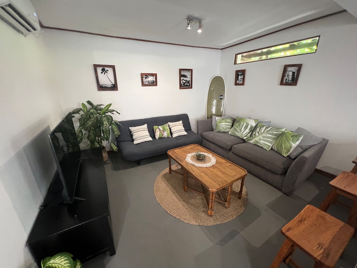 The living room features a comfortable seating area with two sofas adorned with tropical-inspired cushions. A wooden coffee table sits at the center, surrounded by a rug. A television is positioned against one wall, while natural light filters through a small window.