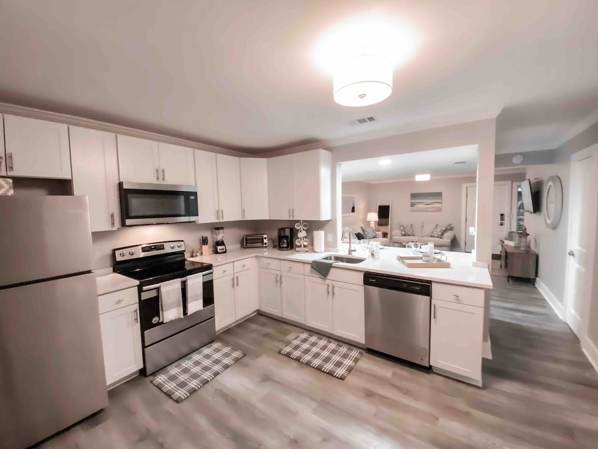 A modern kitchen features stainless steel appliances, including a refrigerator, oven, and dishwasher. White cabinetry complements the light countertops. A double sink is visible, with dishware and small appliances arranged on the countertops. Textured area rugs lie on the wooden floor.