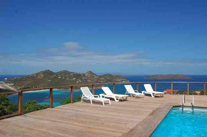A sun-soaked terrace features several white lounge chairs arranged around a sparkling infinity pool. The pool offers stunning views of the blue ocean and surrounding islands, while the wooden deck creates a welcoming and spacious outdoor area.