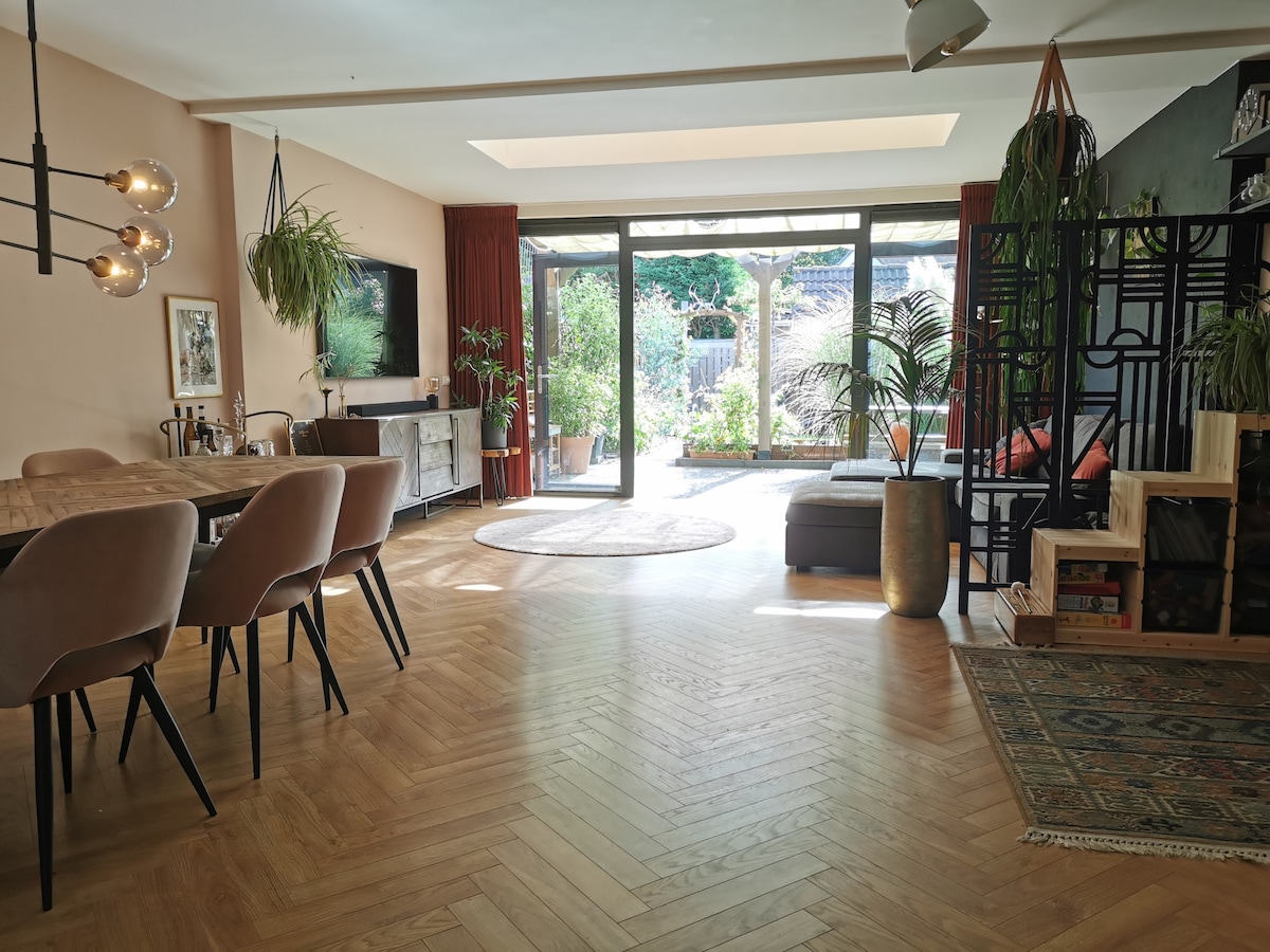 A light-filled living area is showcased, featuring a spacious dining table surrounded by upholstered chairs. Glass doors lead to a garden visible in the background, with greenery enhancing the space. A cozy seating area is present to one side, adorned with plants and decorative elements.