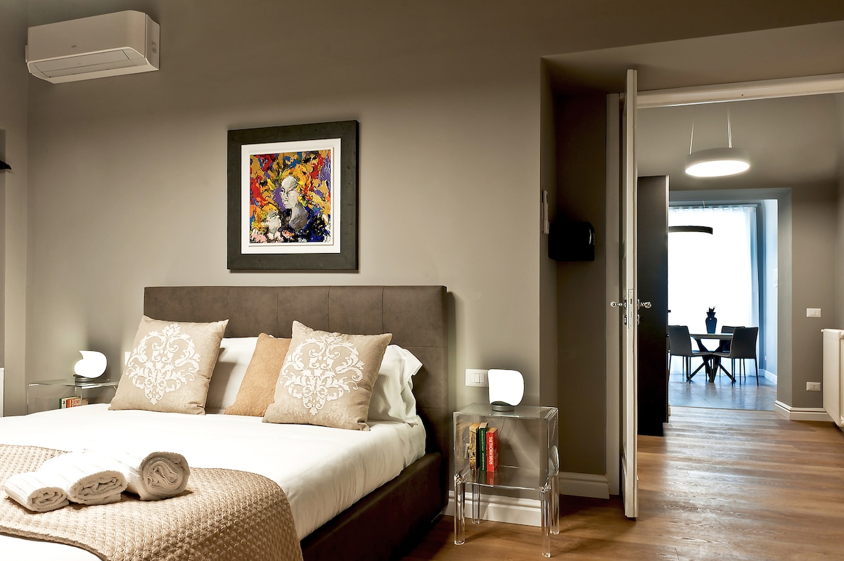 The stylish bedroom is furnished with a spacious bed adorned with decorative pillows and a soft throw. An art piece adds visual interest to the wall above the bed. A small side table holds books, and a door leads to the adjacent dining area with a table and chairs.