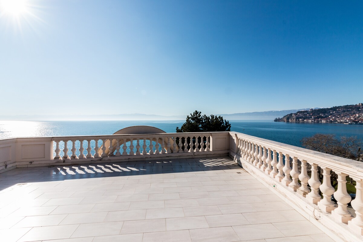 A spacious terrace features a smooth tiled surface, bordered by elegant white balustrades. Expansive views of Ohrid Lake and the distant shoreline create a serene backdrop under a clear blue sky, with the sun casting gentle reflections on the water.
