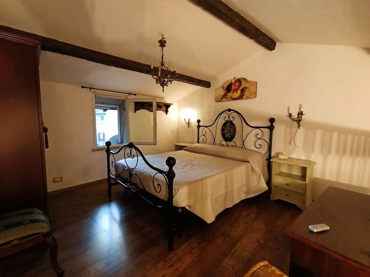 A cozy bedroom is furnished with an elegant wrought-iron bed and complemented by wooden flooring. Natural light enters through a nearby window. A mirror hangs on the wall, while decorative wall sconces and a classic wardrobe enhance the room's charm.