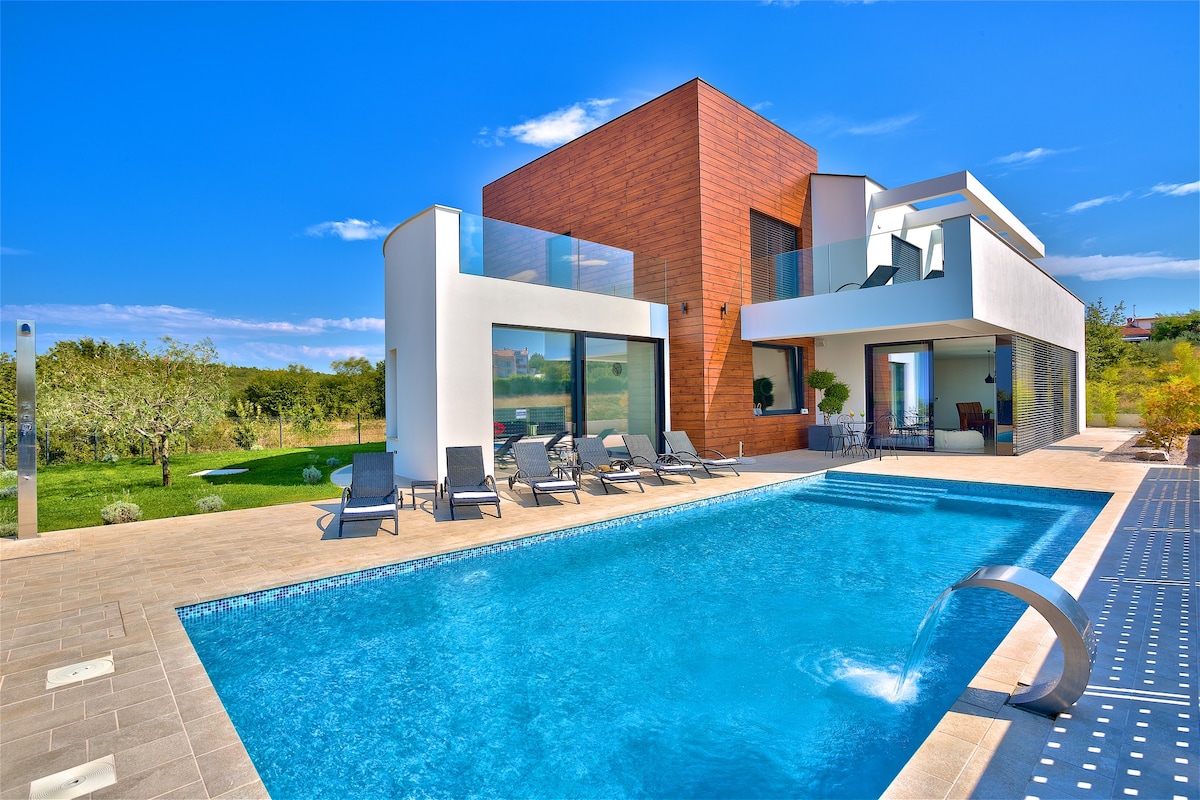 The modern villa features a large swimming pool with a water feature, surrounded by sun loungers. The exterior showcases a combination of white and wooden finishes, with expansive glass doors offering views into the interior. Lush greenery is visible in the background.