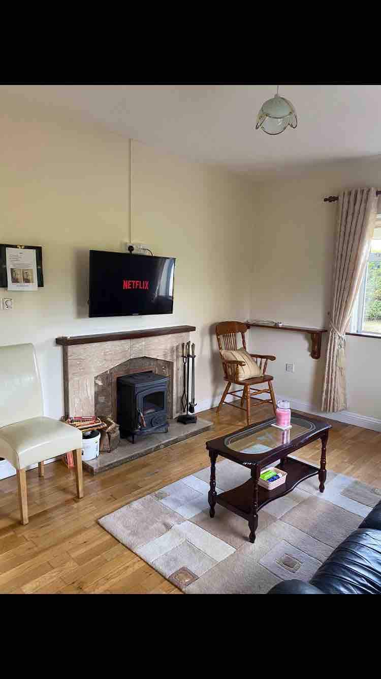 A cozy sitting room is furnished with a comfortable leather sofa and a wooden coffee table. A flat-screen television is mounted on the wall above a classic fireplace, providing a focal point. Natural light filters through the window, enhancing the inviting atmosphere.