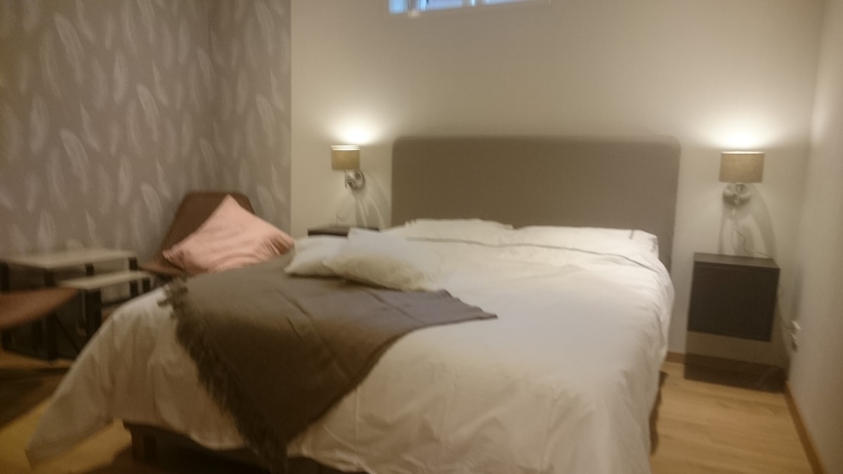 A neatly arranged bedroom features a spacious bed with light bedding and a decorative throw. Two bedside lamps provide soft illumination, while a small side table is positioned next to a comfortable chair. A soft pink cushion enhances the inviting decor.