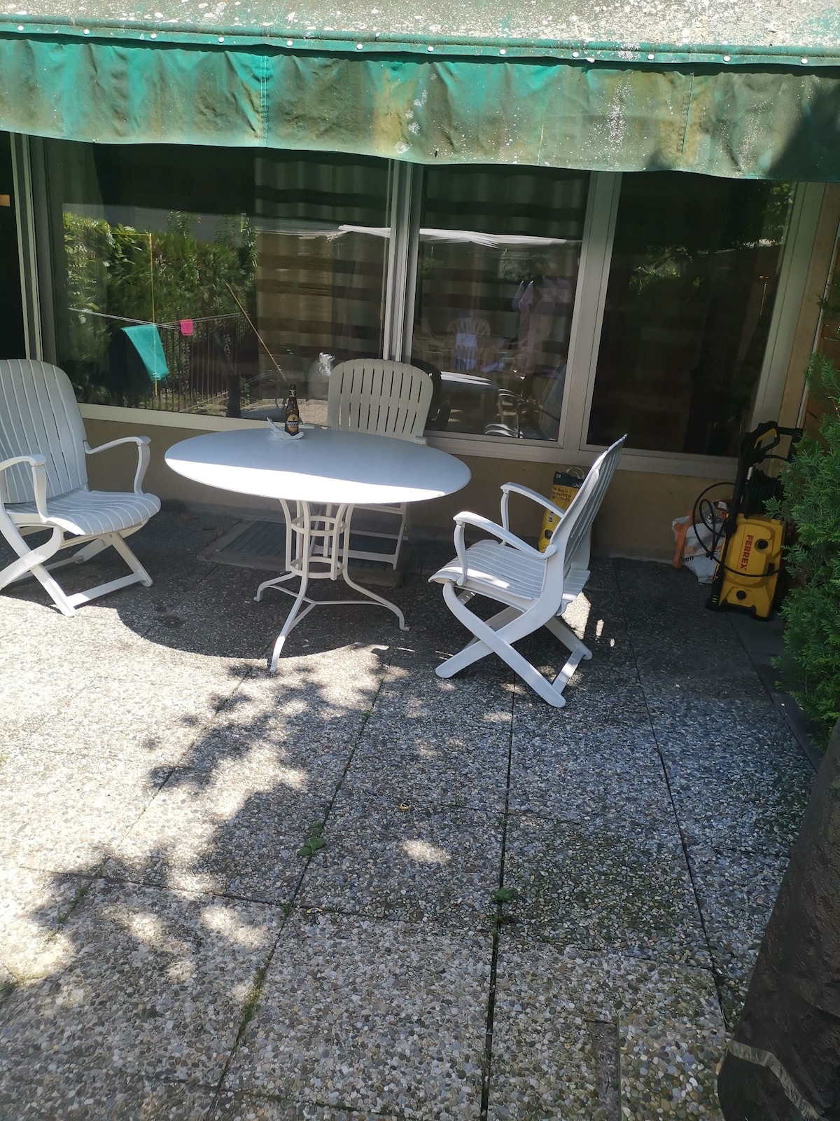 A patio area features a round table surrounded by four white chairs. The space includes a shaded overhang, and sunlight casts shadows on the textured stone flooring. Lush greenery is visible in the background, adding a serene touch to the outdoor setting.