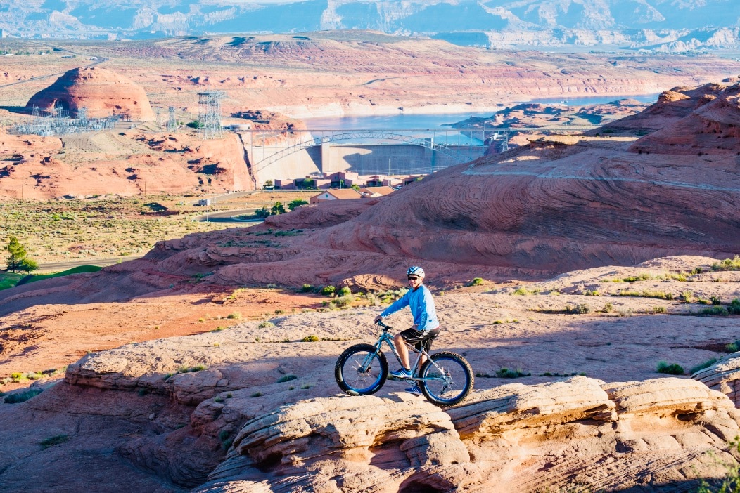 A mountain biker is positioned on a rocky outcrop, overlooking a vast landscape of red rock formations and desert terrain. A reservoir and infrastructure are visible in the distance, framed by layers of mountains under a clear sky.