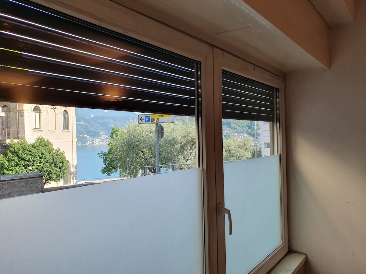 Two windows feature horizontal blinds, offering a view of the lake and surrounding hills. The lower portion of the windows is obscured by frosted glass for privacy. Natural light is filtered into the room, creating a bright ambiance.