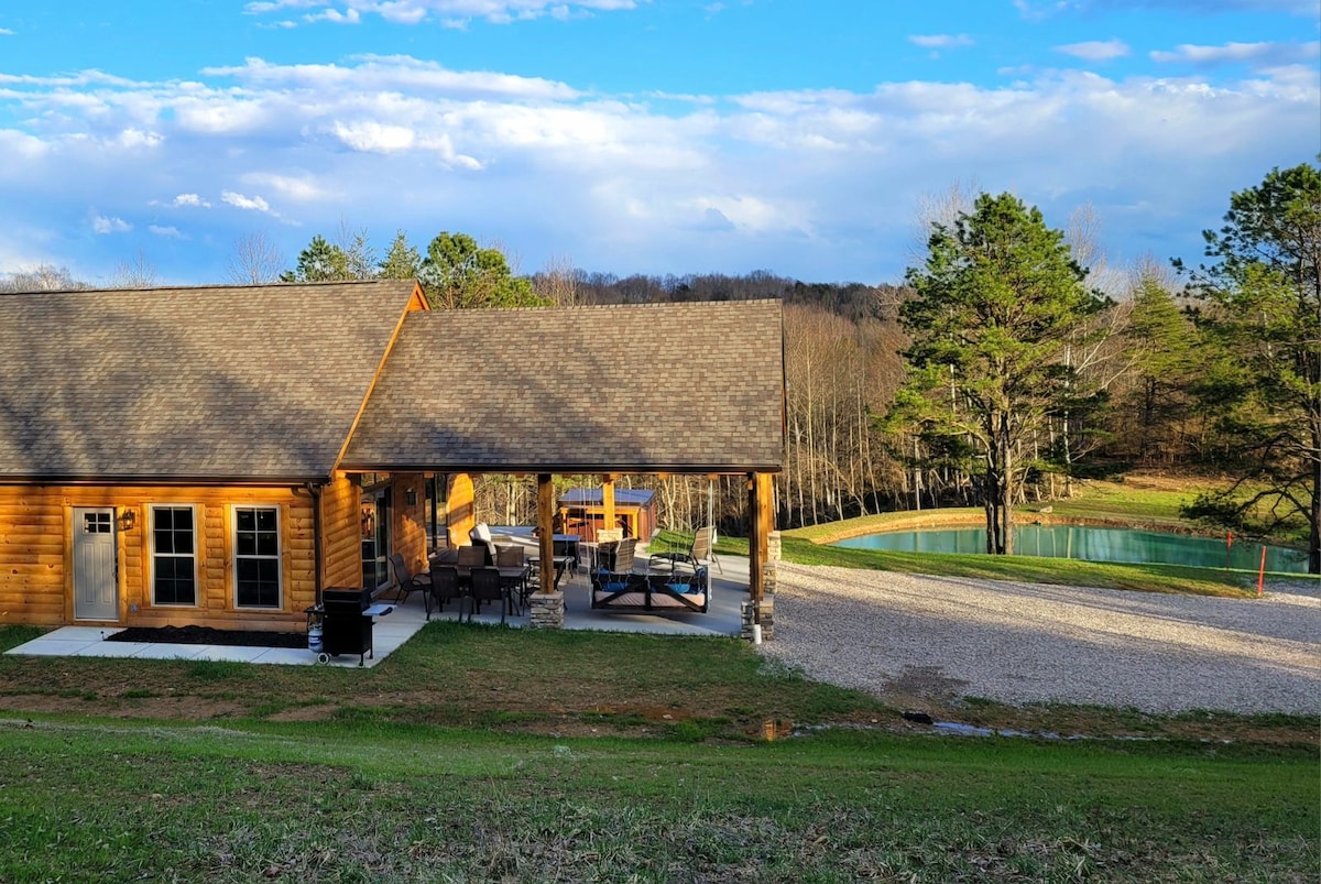 The cabin exterior is showcased, featuring rustic wooden siding and a spacious patio area. A comfortable seating arrangement is visible under the overhang. The background includes a tranquil pond, surrounded by natural greenery and trees, under a partly cloudy sky.