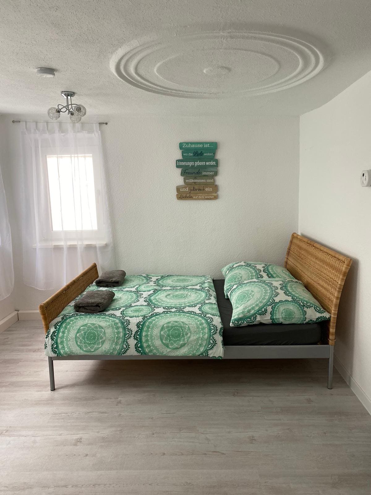 A bright bedroom is furnished with a double bed featuring a patterned comforter in green and white. Two neatly arranged towels are placed on the bed. Natural light filters through a sheer curtain covering the window, while decorative wall signs add a personal touch.
