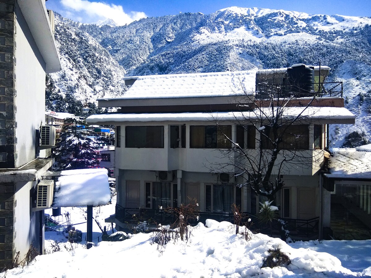 The duplex cottage is surrounded by a blanket of snow, with snow-covered roof and balconies. Majestic snow-capped mountains rise in the background, while a clear blue sky enhances the winter scenery. The garden area is partially visible, featuring a mix of snow and greenery.